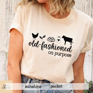 K&ouml;nnte beinhalten: Ein cremefarbenes T-Shirt mit einer schwarzen Grafik einer Henne, einer Pflanze, einem Brotlaib, einem Milchglas und einer Kuh. Der Text "old-fashioned on purpose" befindet sich unter der Grafik.