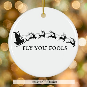 May include: White ceramic Christmas ornament with a black graphic of a witch in a sleigh being pulled by reindeer. The text "FLY YOU FOOLS" is below the graphic. A gold string is attached for hanging.