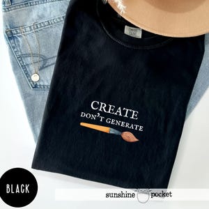 May include: A black t-shirt featuring the white text "CREATE DON'T GENERATE" above an illustration of an orange and brown paintbrush. The shirt has a Comfort Colors label at the inner neckline and is layered over light blue denim fabric.