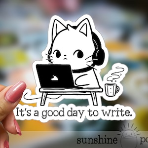 May include: Black and white sticker of a cat wearing headphones and sitting at a desk with a laptop and a cup of coffee. The text "It's a good day to write." is below the cat.