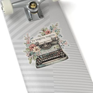 K&ouml;nnte beinhalten: Eine antike Schreibmaschine mit floralen Akzenten auf einem grau-wei&szlig; gestreiften Skateboard-Deck. Die Schreibmaschine hat ein Zitat dar&uuml;ber, das lautet "Um zu beginnen, beginne." und wird William Wordsworth zugeschrieben.