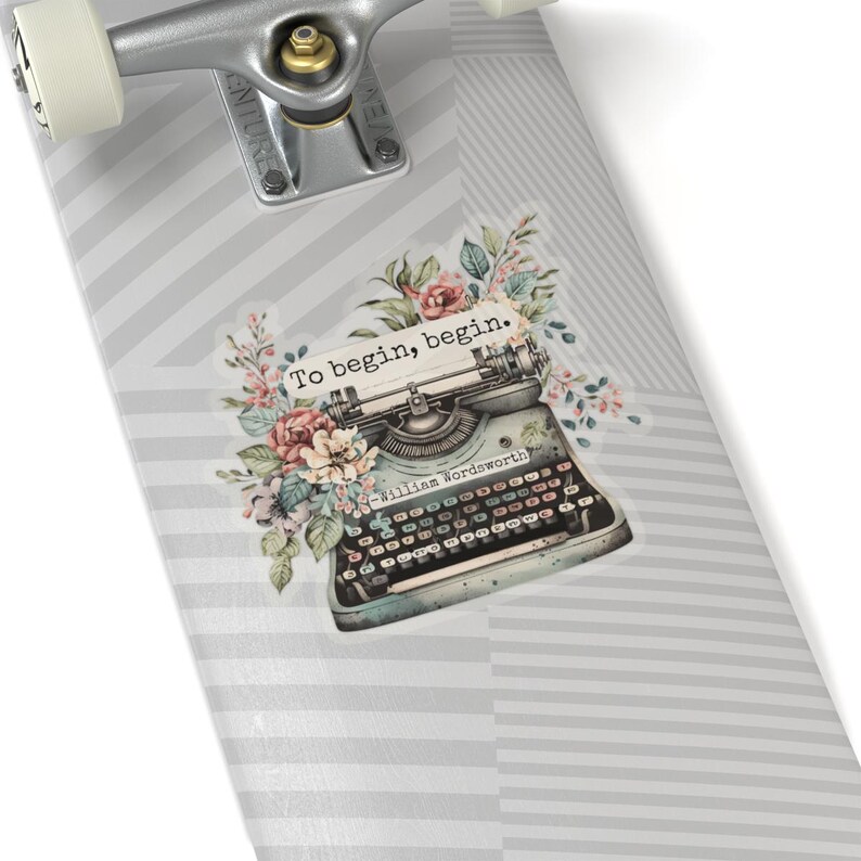 K&ouml;nnte beinhalten: Eine antike Schreibmaschine mit floralen Akzenten auf einem grau-wei&szlig; gestreiften Skateboard-Deck. Die Schreibmaschine hat ein Zitat dar&uuml;ber, das lautet "Um zu beginnen, beginne." und wird William Wordsworth zugeschrieben.