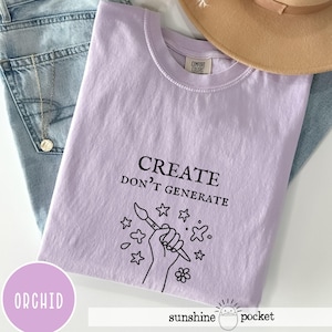 May include: A light purple short-sleeve t-shirt featuring the black text "CREATE DON'T GENERATE" above a line art graphic of a hand holding a paintbrush surrounded by small stars and a flower. The shirt has a crew neckline and a Comfort Colors brand label.
