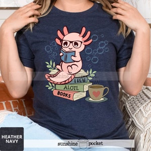 May include: Heather navy t-shirt featuring a cartoon axolotl wearing glasses and reading a book. The axolotl is sitting on a stack of books with the words "I READ ALOTL BOOKS". A coffee cup with a heart is also shown.