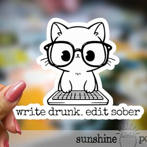 May include: A white cat sticker wearing glasses and sitting at a keyboard. The sticker has the text "write drunk. edit sober." The sticker is held by a hand with pink nail polish. The sticker is from "sunshine pocket."