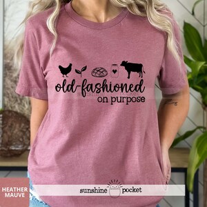 K&ouml;nnte beinhalten: Ein T-Shirt in Heide-Mauve mit einer schwarzen Grafik einer Henne, einer Pflanze, einem Brotlaib, einem Milchglas und einer Kuh. Der Text "old-fashioned on purpose" ist unter der Grafik gedruckt.
