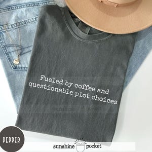 May include: A dark gray t-shirt with the text "Fueled by coffee and questionable plot choices" printed in white.