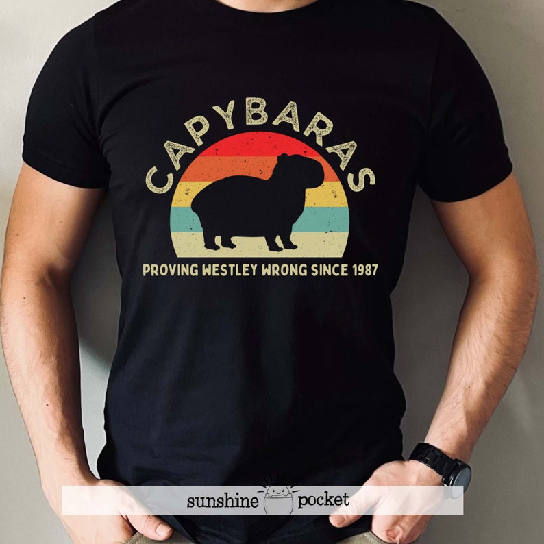 Pu&ograve; includere: Maglietta nera con una grafica in stile vintage di un capibara davanti a un tramonto. Il testo "Capybaras Proving Westley Wrong Since 1987" &egrave; stampato sotto la grafica.