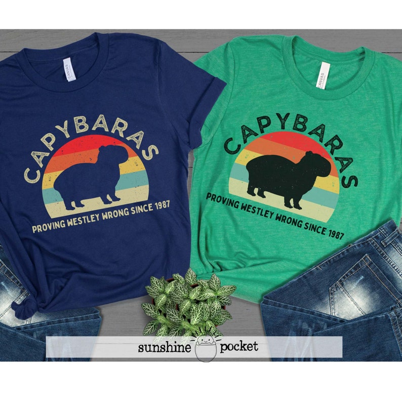 Pu&ograve; includere: Due t-shirt con una grafica di tramonto retr&ograve; e una silhouette di un capibara. Il testo sulle magliette recita "Capybaras Proving Westley Wrong Since 1987".