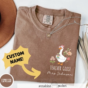 May include: Brown t-shirt featuring a cartoon goose wearing glasses and holding books, with the words "Teacher Goose." The shirt also has the text "Custom Name!" and "Mrs. Johnson."