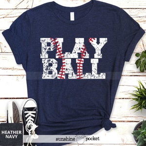 May include: A navy blue t-shirt with a white distressed graphic that says "PLAY BALL" with a red baseball stitched into the graphic.