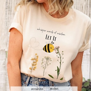 May include: A beige t-shirt with a bee graphic and the text "whisper words of wisdom LET IT". The bee is yellow and black and is flying towards a cluster of dried flowers.