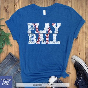 May include: A blue t-shirt with a white distressed graphic that says "PLAY BALL" with a baseball graphic in the middle of the word "PLAY".