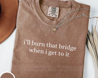 May include: Espresso-colored Comfort Colors t-shirt with the phrase "I'll burn that bridge when I get to it" in white. A silver necklace with a pendant rests on the shirt. The shirt has a crew neck and short sleeves.