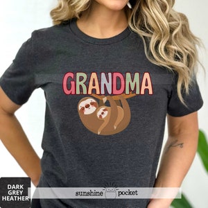 May include: Dark heather grey t-shirt with a graphic of two sloths hanging from a branch. The text "Grandma" is above the sloths in colorful letters.