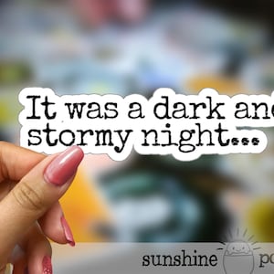 May include: A white sticker with the text "It was a dark and stormy night..." in a bold, black font. The sticker is held by a hand with pink nail polish. The background is blurred with various colors.