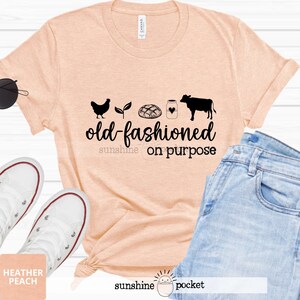 K&ouml;nnte beinhalten: Ein T-Shirt in Pfirsichfarbe mit einer schwarzen Grafik einer Henne, einer Pflanze, einem Brotlaib, einem Milchglas, einem Herzen und einer Kuh. Der Text "old-fashioned sunshine on purpose" ist unter der Grafik gedruckt.