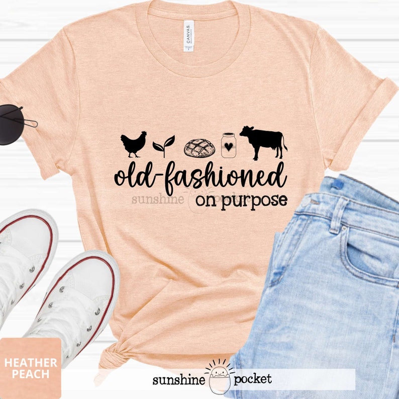 K&ouml;nnte beinhalten: Ein T-Shirt in Pfirsichfarbe mit einer schwarzen Grafik einer Henne, einer Pflanze, einem Brotlaib, einem Milchglas, einem Herzen und einer Kuh. Der Text "old-fashioned sunshine on purpose" ist unter der Grafik gedruckt.