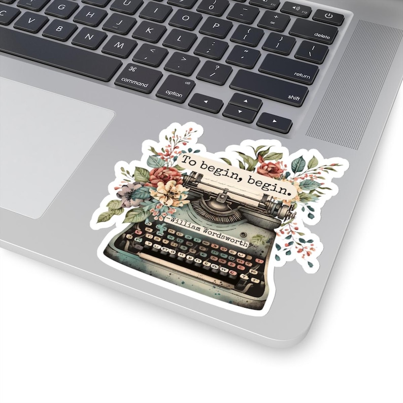 K&ouml;nnte beinhalten: Eine Vintage-Schreibmaschine mit floralen Akzenten und dem Zitat "Um zu beginnen, beginne." von William Wordsworth. Die Schreibmaschine ist blau und hat einen wei&szlig;en Papiereinsatz.