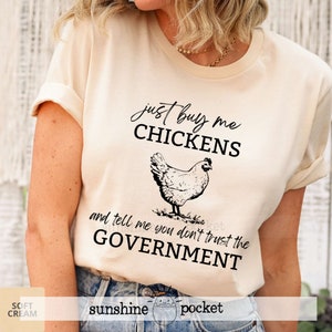 May include: A beige t-shirt with a black graphic of a chicken and the text "just buy me chickens and tell me you don't trust the government".