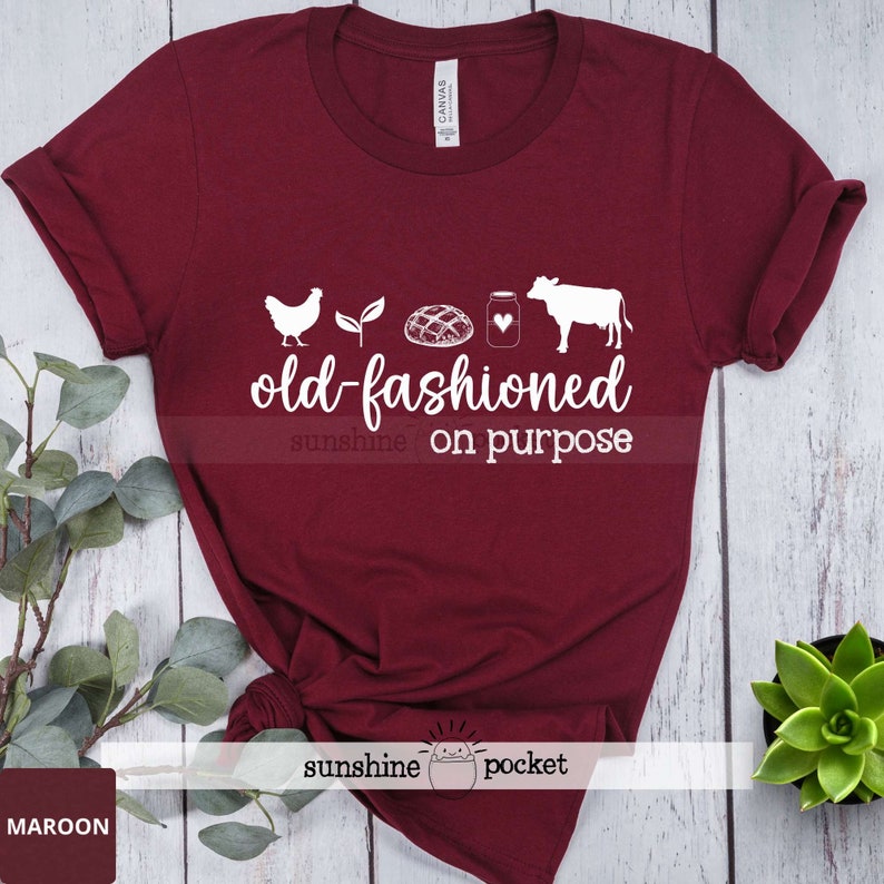 K&ouml;nnte beinhalten: Maroonfarbenes T-Shirt mit wei&szlig;em Text, der "old-fashioned sunshine on purpose" lautet. Der Text wird von einem Huhn, einer Pflanze, einem Brotlaib, einem Milchglas und einer Kuh begleitet.