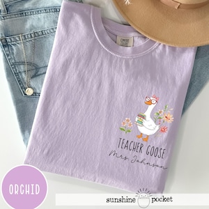 May include: Orchid-coloured t-shirt featuring a goose wearing glasses and holding books, surrounded by flowers. The text "TEACHER GOOSE Mrs. Johnson" is printed below the goose. The shirt is folded on top of blue jeans.