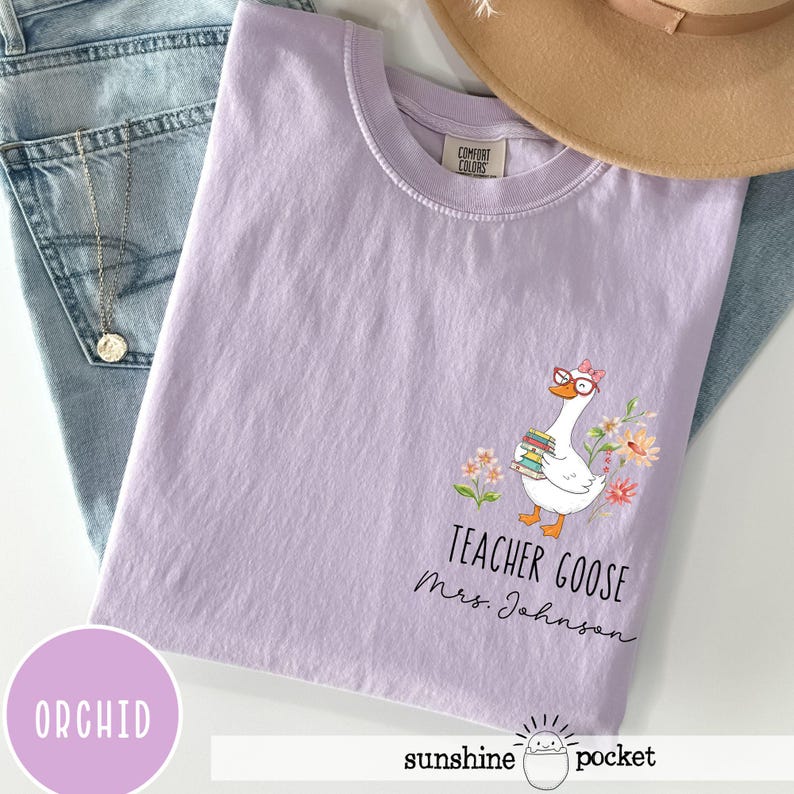 May include: Orchid-coloured t-shirt featuring a goose wearing glasses and holding books, surrounded by flowers. The text "TEACHER GOOSE Mrs. Johnson" is printed below the goose. The shirt is folded on top of blue jeans.