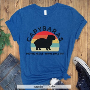 Pu&ograve; includere: Una maglietta blu con una grafica in stile vintage di un capibara davanti a un tramonto. Il testo recita "Capybaras Proving Westley Wrong Since 1987".