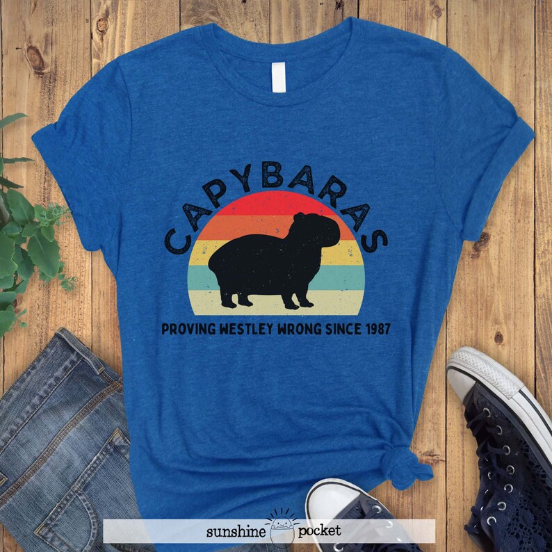 Pu&ograve; includere: Una maglietta blu con una grafica in stile vintage di un capibara davanti a un tramonto. Il testo recita "Capybaras Proving Westley Wrong Since 1987".