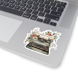K&ouml;nnte beinhalten: Eine Vintage-Schreibmaschine mit floralen Akzenten und dem Zitat "Um zu beginnen, beginne." von William Wordsworth. Die Schreibmaschine ist blau und wei&szlig; mit einer schwarzen Tastatur. Die Blumen sind rosa, rot und gelb.