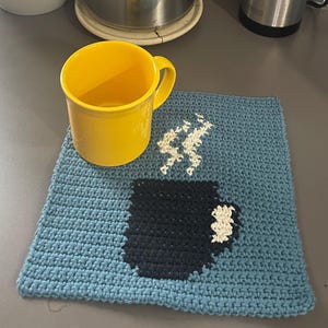 May include: A yellow mug sits next to a blue crocheted coaster with a black coffee cup design and white steam. A stainless steel French press, coffee grinder, and potted plant are in the background.