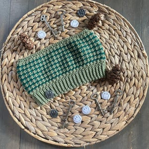 Może przedstawiać: Zielona i ciemnozielona czapka typu beanie z szydełka z prążkowanym brzegiem, spoczywająca na okrągłej, tkanej podkładce. Scena obejmuje zabytkowe klucze, małe białe guziki z czarną grafiką i szyszki.