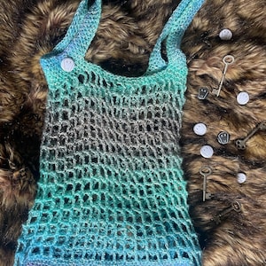 May include: A handmade crochet tote bag in shades of blue, teal, and purple. The open-weave design features a button closure and sturdy straps. Decorative keys and small round pins with skull and crossbones are scattered around the bag.