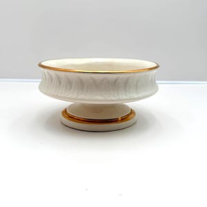May include: A small, off-white pedestal bowl with a gold rim and base trim. The bowl has a textured, ribbed design around the sides. The pedestal base is round and the bowl is likely for decorative purposes.