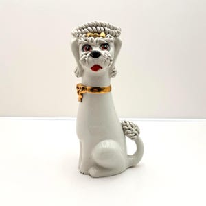 May include: A white ceramic poodle figurine with a gold collar and accents. The poodle has red eyes, a black nose, and a red mouth. The figurine has a braided headpiece and a curly tail. The poodle is sitting upright.