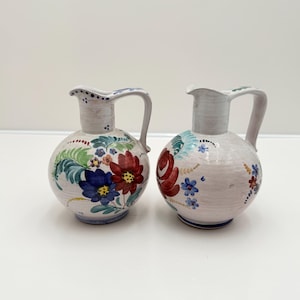 May include: Two white ceramic pitchers with floral designs. One pitcher features red, blue, and yellow flowers with green leaves. The other pitcher has red and blue flowers with green leaves. Both have handles and are on a white background.