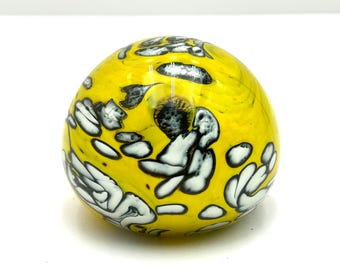 May include: A round, decorative glass paperweight. The paperweight is primarily yellow with a pattern of white and black swirls and shapes throughout. The glass is smooth and glossy.