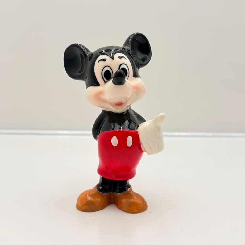 Pu&ograve; includere: Una statuetta in ceramica di Topolino, un classico personaggio Disney. La figura ha testa e orecchie nere, viso bianco e pantaloncini rossi. Mickey fa il pollice in su con una mano guantata bianca. La statuetta &egrave; su una superficie bianca.