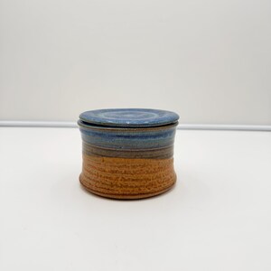 May include: A cylindrical ceramic jar with a matching lid. The upper portion features a smooth, mottled blue glaze, while the lower section displays a textured, earthy orange-brown finish with horizontal ridges circling the base of the container.