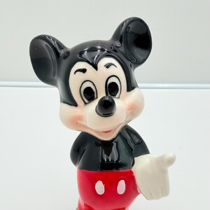 Pu&ograve; includere: Statuetta in ceramica di Topolino. Il corpo e le orecchie sono neri, i pantaloncini rossi con bottoni bianchi, il viso bianco, le guance rosa e il naso nero. La mano destra indica.