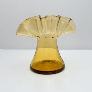 May include: A translucent amber-colored glass vase featuring a wide, flared opening with a distinctively ruffled, wavy rim. The body of the vase tapers inward from the top toward a circular, flat base, showcasing a smooth, polished finish.