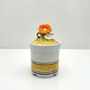 May include: A white ceramic jar with a yellow and brown stripe design. The lid is decorated with a three-dimensional orange and white flower.