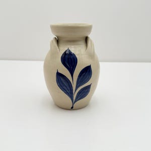 May include: A small, beige ceramic vase with a rounded body and a flared top. The vase features two small handles and is decorated with a blue leaf design. The vase is approximately 5 inches tall.