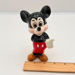 Pu&ograve; includere: Una statuetta in ceramica di Topolino. La figura indossa una giacca nera, pantaloncini rossi con due bottoni bianchi e scarpe marroni. Mickey ha una mano guantata bianca che indica verso destra. La statuetta ha orecchie nere e un viso bianco con un naso nero.