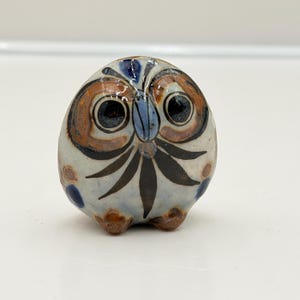 May include: A small, round, hand-painted ceramic owl figurine with a cream-colored base. It features large, dark, circular eyes, a blue beak, and brown and black painted details including leaf-like patterns on the chest and blue spots.