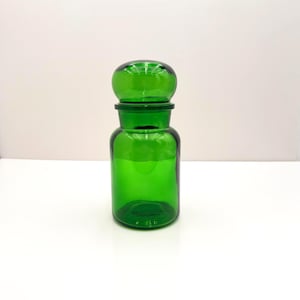 May include: A translucent emerald green glass apothecary jar with a cylindrical body and a rounded, dome-shaped lid. The container features a smooth, polished surface and a wide neck, showcasing a vibrant, uniform green color throughout the glass.