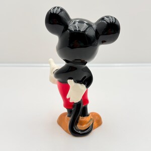 Pu&ograve; includere: Una statuetta in ceramica di Topolino, vista da dietro. La figura ha una testa e orecchie nere lucide, un corpo nero, pantaloncini rossi, guanti bianchi e scarpe arancioni. La statuetta &egrave; su una superficie bianca.