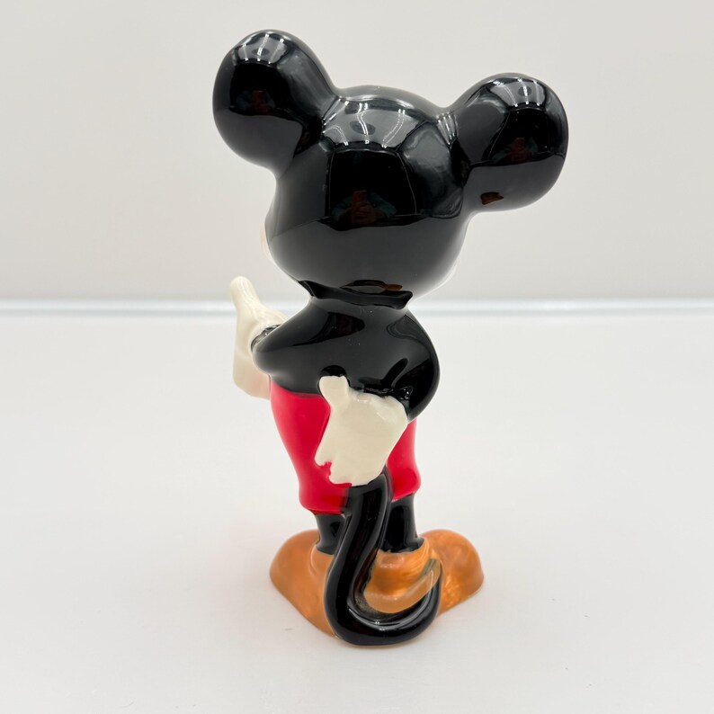 Pu&ograve; includere: Una statuetta in ceramica di Topolino, vista da dietro. La figura ha una testa e orecchie nere lucide, un corpo nero, pantaloncini rossi, guanti bianchi e scarpe arancioni. La statuetta &egrave; su una superficie bianca.