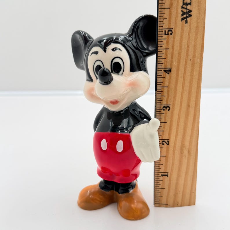 Pu&ograve; includere: Una statuetta in ceramica di Topolino. La figura ha testa e orecchie nere, viso bianco e busto rosso con due bottoni bianchi. I piedi sono marroni e le mani bianche. Un righello di legno &egrave; accanto alla statuetta.