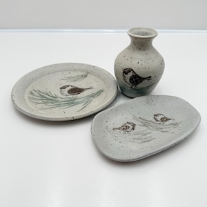 May include: A ceramic set featuring a small vase and two plates, all with a speckled, light gray finish. Each piece is adorned with a hand-painted bird and foliage design in brown and green. The set is ideal for decorative use.